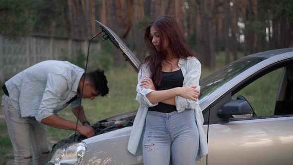 Bored Dissatisfied Girlfriend Leaning on Car in Forest Talking to Man Repairing Broken Vehicle alt