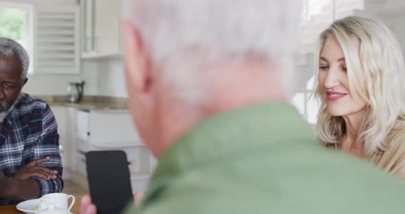 Two diverse senior couples sitting by a table drinking tea using smartphones at home alt
