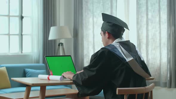 Asian Man Wearing A Graduation Gown And Cap Use Laptop Computer With Green Mock Up Screen alt