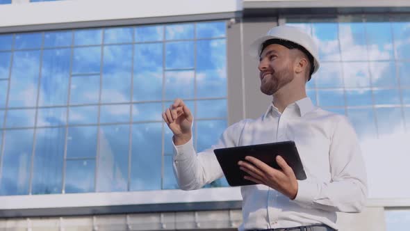 An Engineer Architect in a White Shirt and Helmet on the Background of a Modern Glass Building Works alt