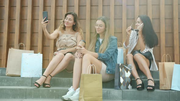 Joyful Women Friends Using Smart Phones and Drinking to Go Coffee Sitting Outdoors with Shopping alt