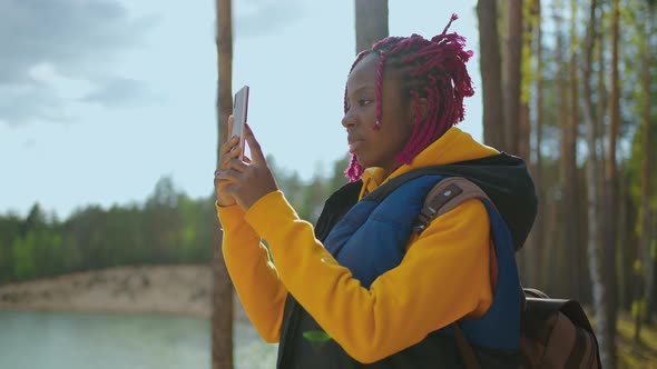 A Black Girl with a Backpack Uses a Mobile Phone While Hiking Through the Forest in the Mountains alt