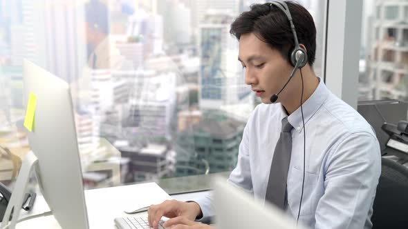 Young Asian man telemarketing agent wearing microphone headset typing on computer keyboard
