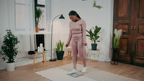 Young Happy African American Woman Standing on Scales and Enjoying Weight Loss Checking Slimming at alt