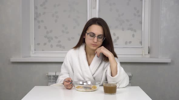 Sleepy Young Woman in a White Robe Eats Cereal Drinks Coffee and Invigorates alt