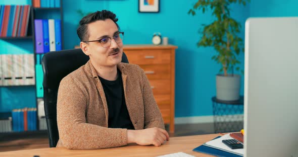 A Man Wearing Glasses with a Mustache Sits in a Company Office in Front of alt