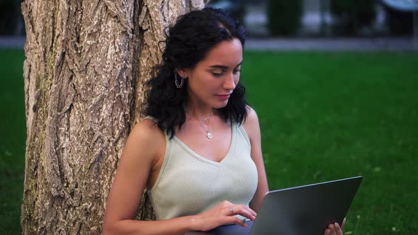 Portrait of a Woman in Dress Sits Below Tree at the Park on Grass Holding Laptop on Her Knees Typing alt