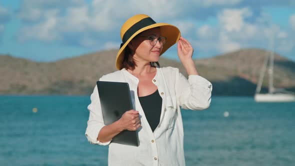 Outdoor Portrait of Mature Woman in Hat on Beach with a Laptop in Hands alt