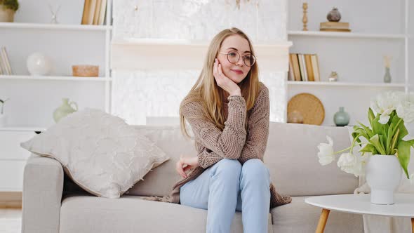 Happy Young Caucasian Mom Blonde Woman in Glasses Nanny Sits on Sofa in Living Room Looks at Little alt
