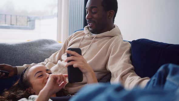 Relaxed Young Couple At Home On Sofa Watching TV And Checking Social Media On Mobile Phone