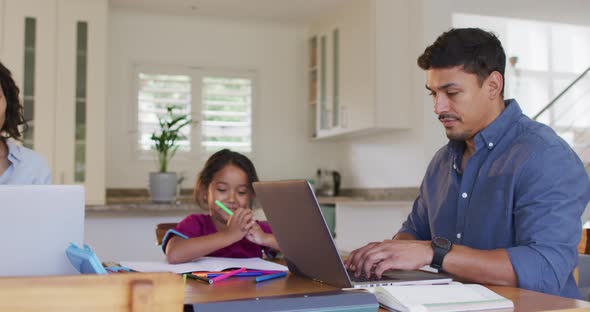 Hispanic parents sitting at table working with laptops and daughter drawing alt