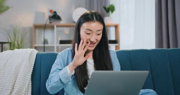 Asian Brunette Sitting on the Couch at Home and Waving Hand to Greet Her Interlocutor Via Video Call alt