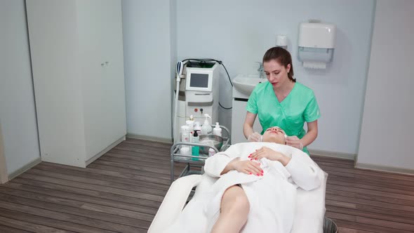Girl Beautician Makes Facial Cleansing Procedure for Young Woman in Beauty Salon alt