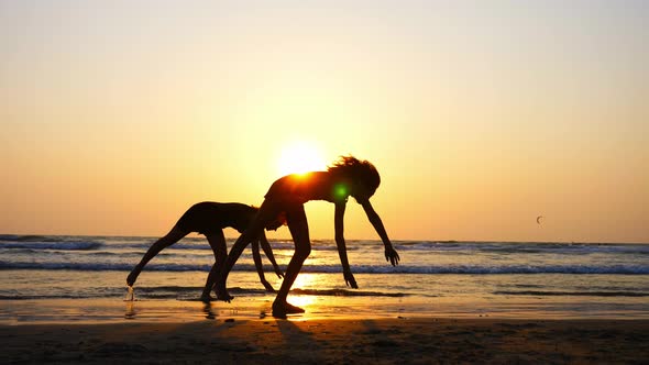 Silhouette of Sporty Young Women Practicing Acrobatic Element on the Beach alt