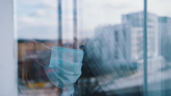 Portrait of Lonely Elderly Woman in Quarantine with Face Mask Looking Through the Window alt