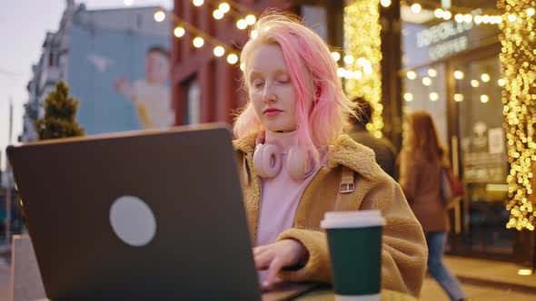 Young Pink Haired Stylish Woman Working on Laptop Sitting Outdoors with Takeout Coffee and Computer alt