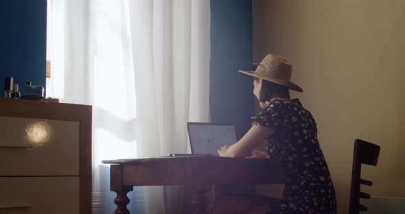 Girl in Hat Sitting at Table with Laptop Working at Home By Window