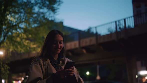Young woman smiling while typing into her phone at night alt