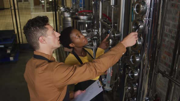 Diverse male and female colleague at gin distillery inspecting equipment and discussing alt