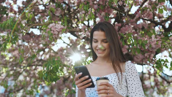 Attractive Woman With Smartphone and Cup of Coffee in Hands on Background of Sakura Tree in City