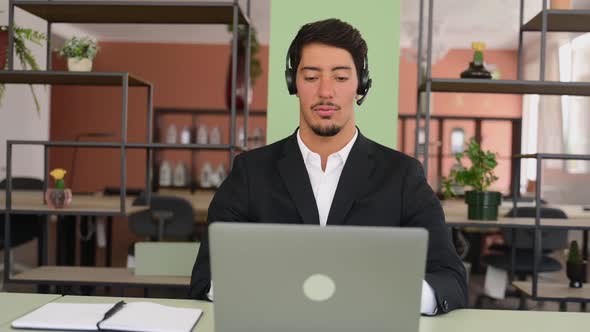 Smiling Latin Male Employee Wearing Headset Talking Online Sitting Near Windows in the Office