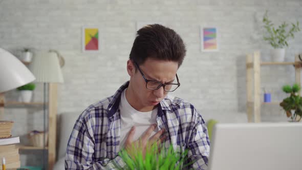 Young Asian Man in a Shirt Coughs Sitting at the Laptop alt
