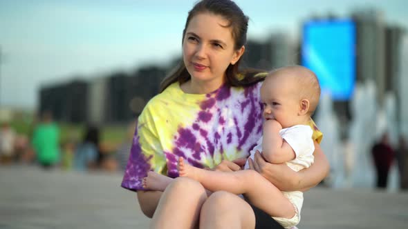 Modern Mother Holds Ten Month Old Son in Her Arms and Sit on the Sidewalk Relax Outdoors in the Park