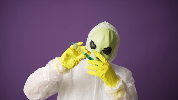 A Man in an Alien Mask in a White Suit Shows a Syringe with a Covid Vaccine on a Purple Background alt