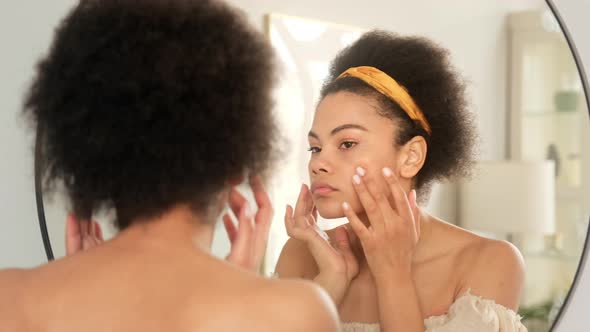 Young african american woman looking touching  face in the mirror examining skin condition alt