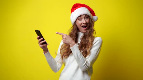 Woman Christmas Santa Hat White Sweater Yellow Studio Background with Smartphone in Hand alt