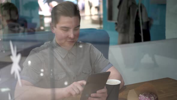 A Young Man is Sitting Behind a Glass in a Cafe with a Tablet in His Hands alt
