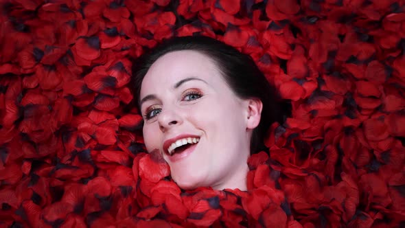 An attractive young woman laying on the floor looking happy ith red rose petals all around her head