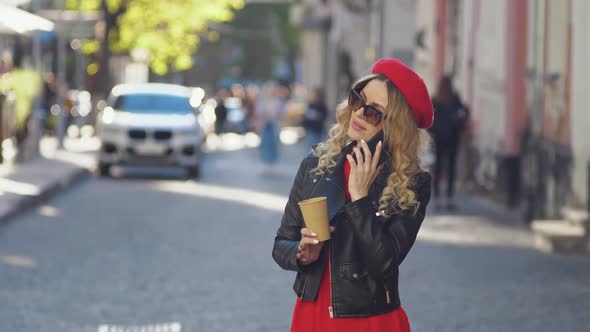 A Young Woman in the Middle of the Street Answers a Cell Phone Call and Drinks Takeaway Coffee alt
