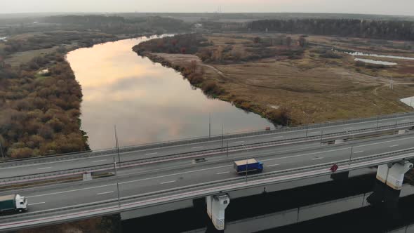 Aerial: Four-lane Road Bridge Over a River in Late Autumn with Two Trucks in Motion alt