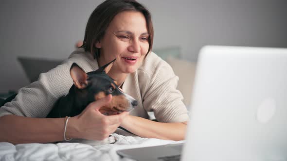 A Woman Is Making a Videocall on Her Laptop While Lying on the Bed with Her Dog alt