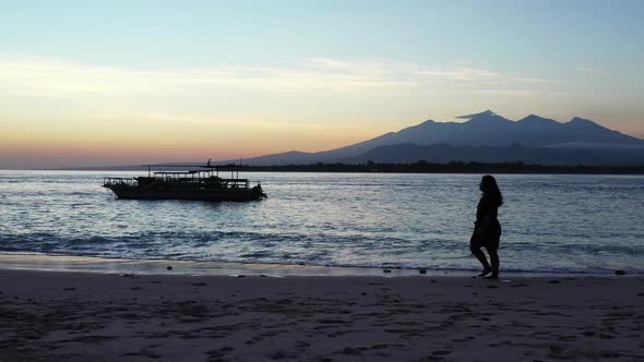 Silhouette of girl walking alone on exotic beach after sunset with bright sky reflecting on calm sea alt