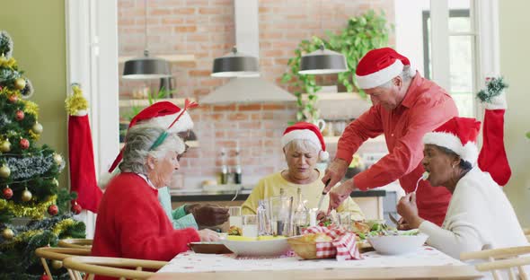 Caucasian senior man in santa hat carving turkey at christmas dinner table with diverse friends alt