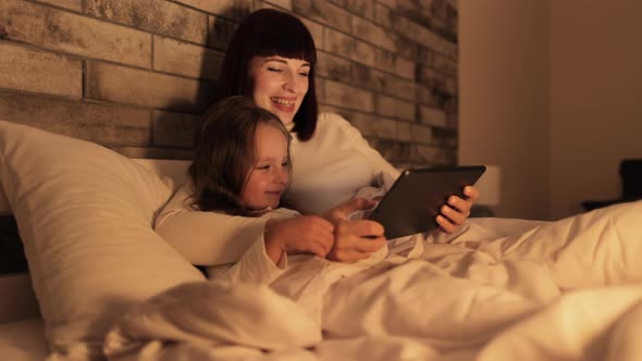 Mother and Daughter Using Tablet Pc Lying in Bed Under the Blanket and Spending Their Evening Time alt