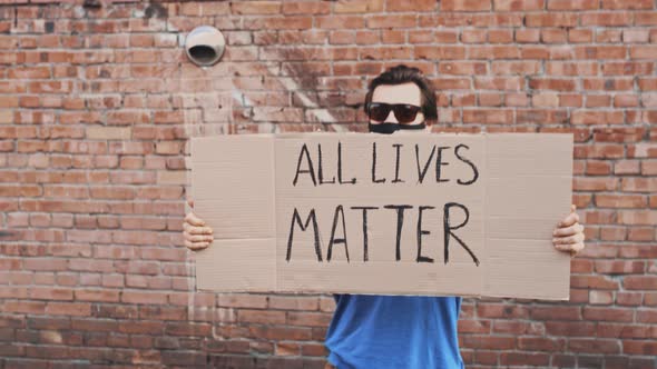 Man in Mask Stands Against Red Wall with Cardboard Poster  ALL LIVES MATTER