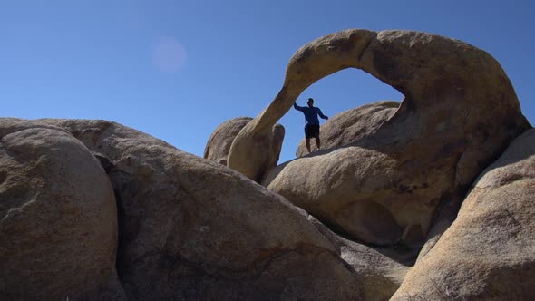 A young man trail running with his dog past a natural rock arch in a mountainous desert. alt