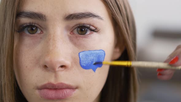 Close-up Portrait of Young Pleasant Woman Looking at Camera While Artist Painting with White on Blue alt