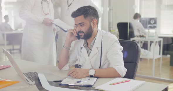 Male Doctor Talking on Phone with Patient and Working at Laptop alt