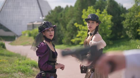 Scared Beautiful Ladies in Steampunk Outfit Looking at Blurred Gun in Hand at Front with Frightened alt