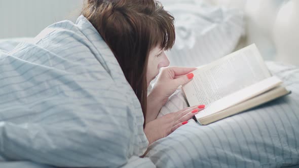 Woman Lies on the Bed and Reads a Book Covered with a Blanket Woman Spends the Day Reading a Book