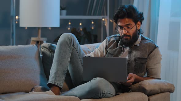 Freelancer Guy Arab Indian Man Bearded Male User Wearing Glasses Sitting on Sofa at Night Late Time alt