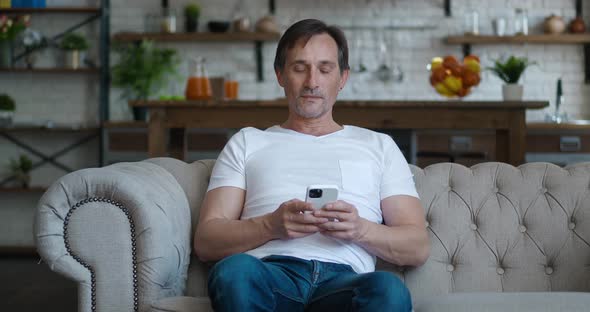 Handsome Caucasian Mature Man Sitting on Sofa at Cosy Home and Using Smartphone Surfing Internet alt