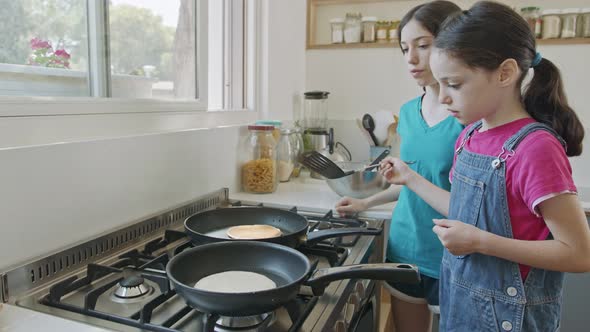 Two young girls preparing pancakes in the kitchen using a frying pan alt