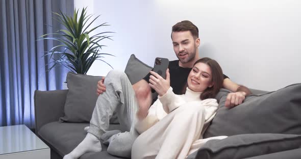 Young Couple Takes a Selfie on a Smartphone a Guy and a Girl Sit on the Sofa in the Living Room and alt