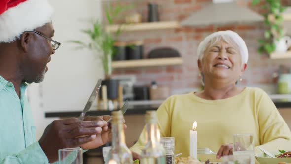 Happy diverse senior friends in santa hats talking and laughing at christmas dinner table at home alt