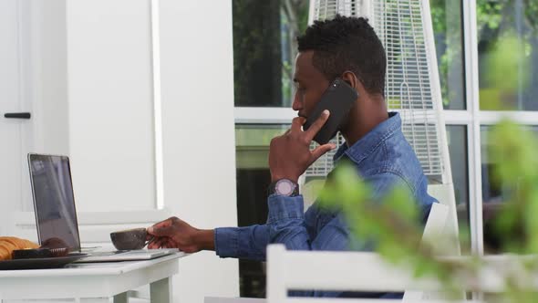 African american businessman using laptop and talking on smartphone in cafe alt